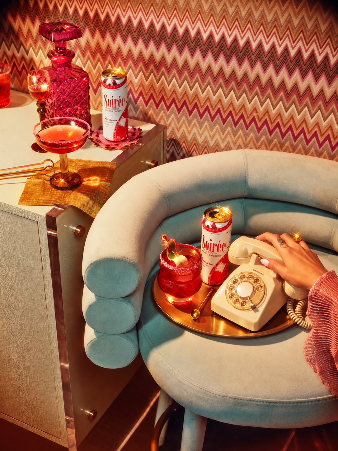 A hand reaching for a vintage cream rotary phone beside Soirée Marge-a-rita cans and cocktails